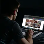 Молодой человек в темноте читает сайт Парни+ на экране ноутбука