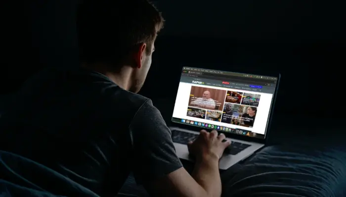 Молодой человек в темноте читает сайт Парни+ на экране ноутбука