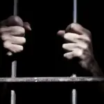 мужские руки держат прутья решётки, иллюстрируют заключение, в камере темно