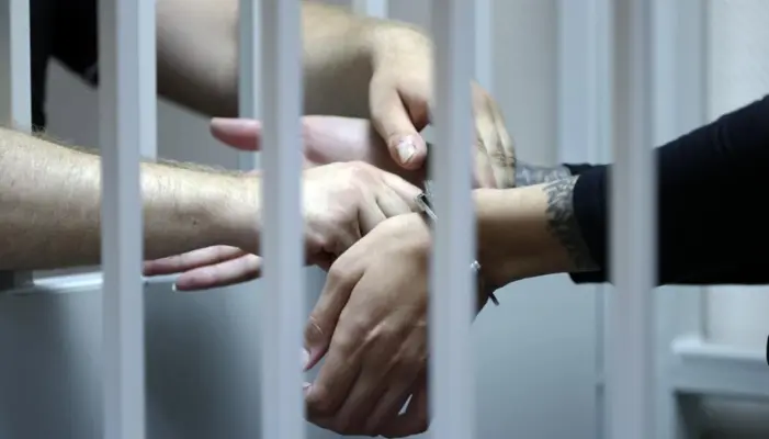 за решёткой видны руки в наручниках и руки полицейского