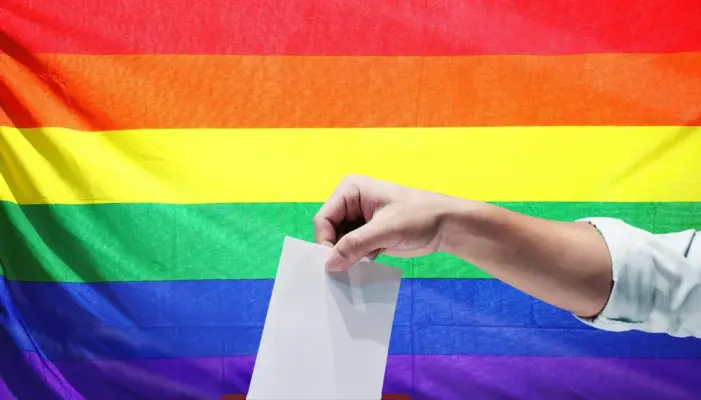 Рука опускает лист в урну на фоне радужного флага. ЛГБТ, голосование, выборы